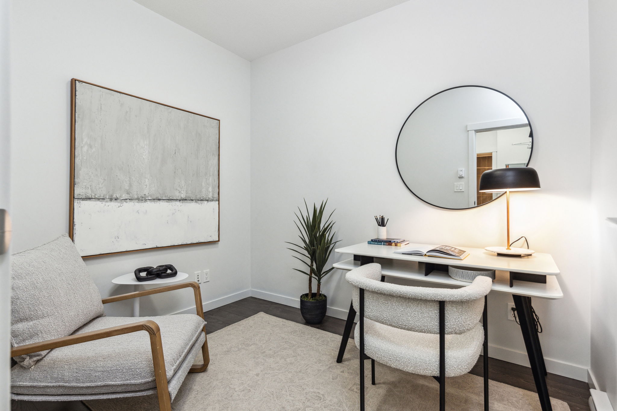 Office with grey painting, mirror, arm chair, desk chair, and desk with lamp.